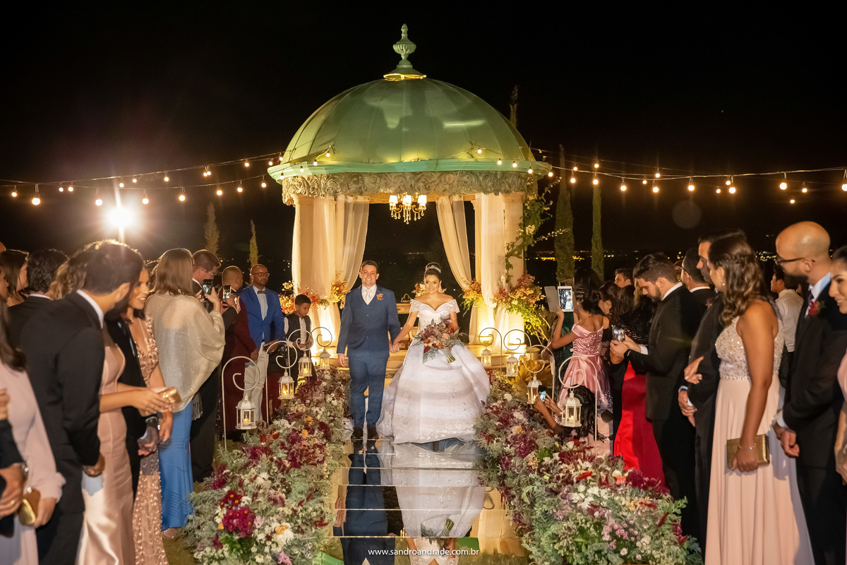 Saída dos noivos, passou nervosismo, o casal está extremamente feliz e cumprimentando todos os convidados, o gazebo ao fundo e um lindo tapete espelhado compõem esta fotografia colorida.
