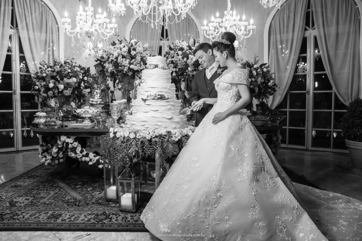 O casal está na frente da mesa do bolo, ainda durante o corte, uma fotografia preto e branca, pegando toda a decoração dessa área da mesa do bolo e dos doces.