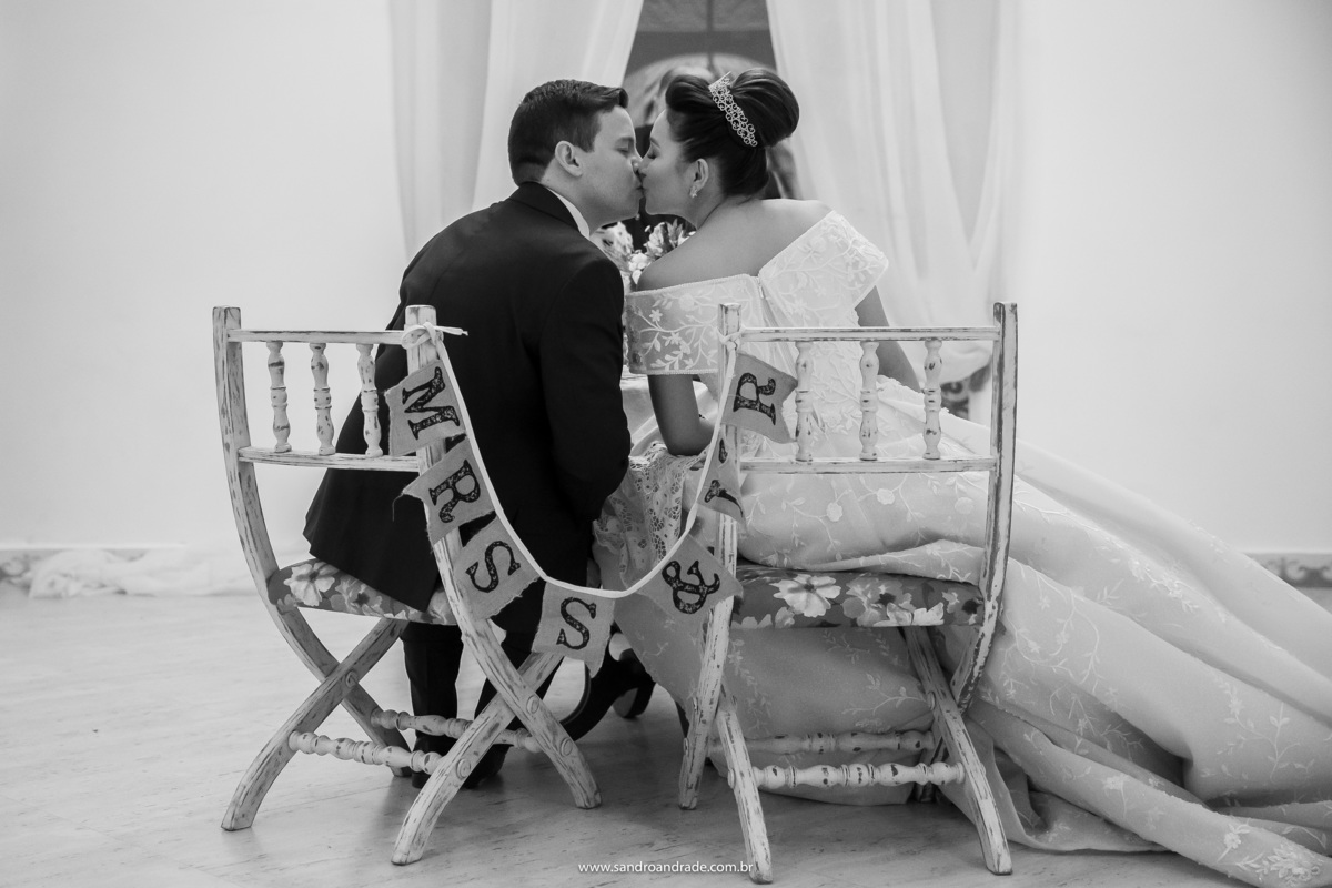 Um beijinho, de costas, sentados nas cadeiras com plaquinhas identificando Senhor e Senhora, durante o jantar a dois.