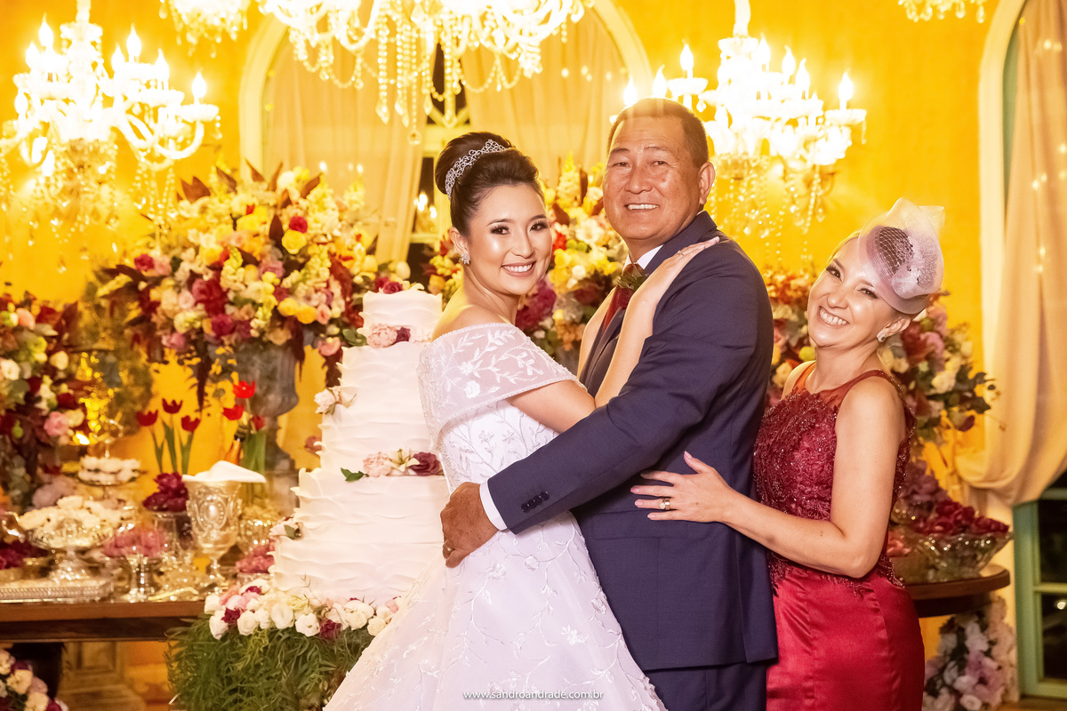 Dany está abraçado com os pais dela na mesa do bolo, todos sorriem para a foto.