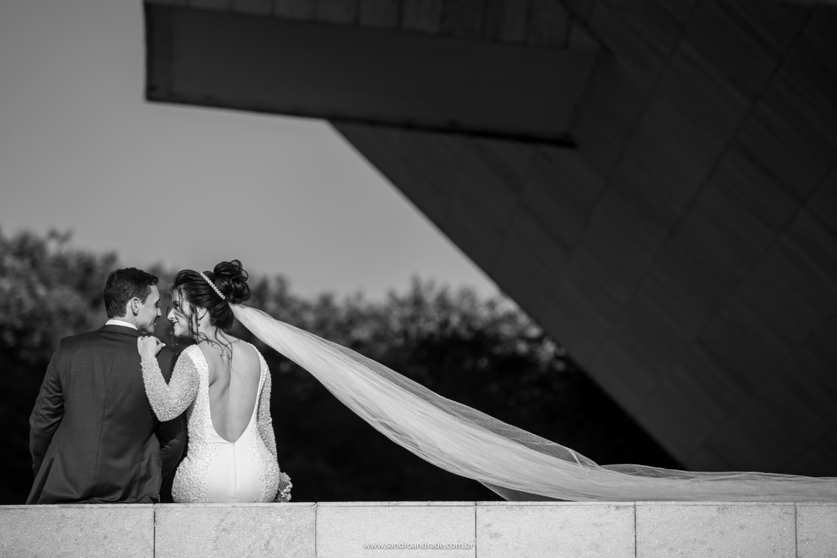 Os noivos estão sentados no muro do panteão da pátria, de costas, parte da arquitetura do prédio em silhueta, um olho no olho e muito amor.