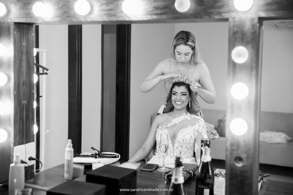 A noiva de frente ao espelho arrumando o cabelo para o grande dia, com uma belíssima coroa de princesa.