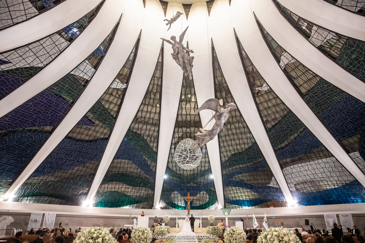 Uma peculiaridade da Catedral de Brasília são os enormes anjos pendurados no teto, esta fotografia mostra toda a igreja e os noivos de costas no altar com toda a grandiosa arquitetura e os anjos. Fotografia e arquitetura uma combinação