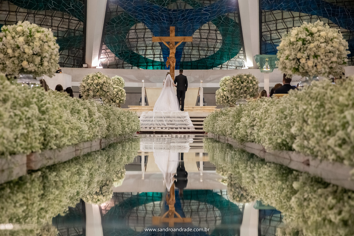 Mais uma fotografia de costas, pegando todo o altar  e os noivos no centro dela com um belo reflexo no tapete espelhado.