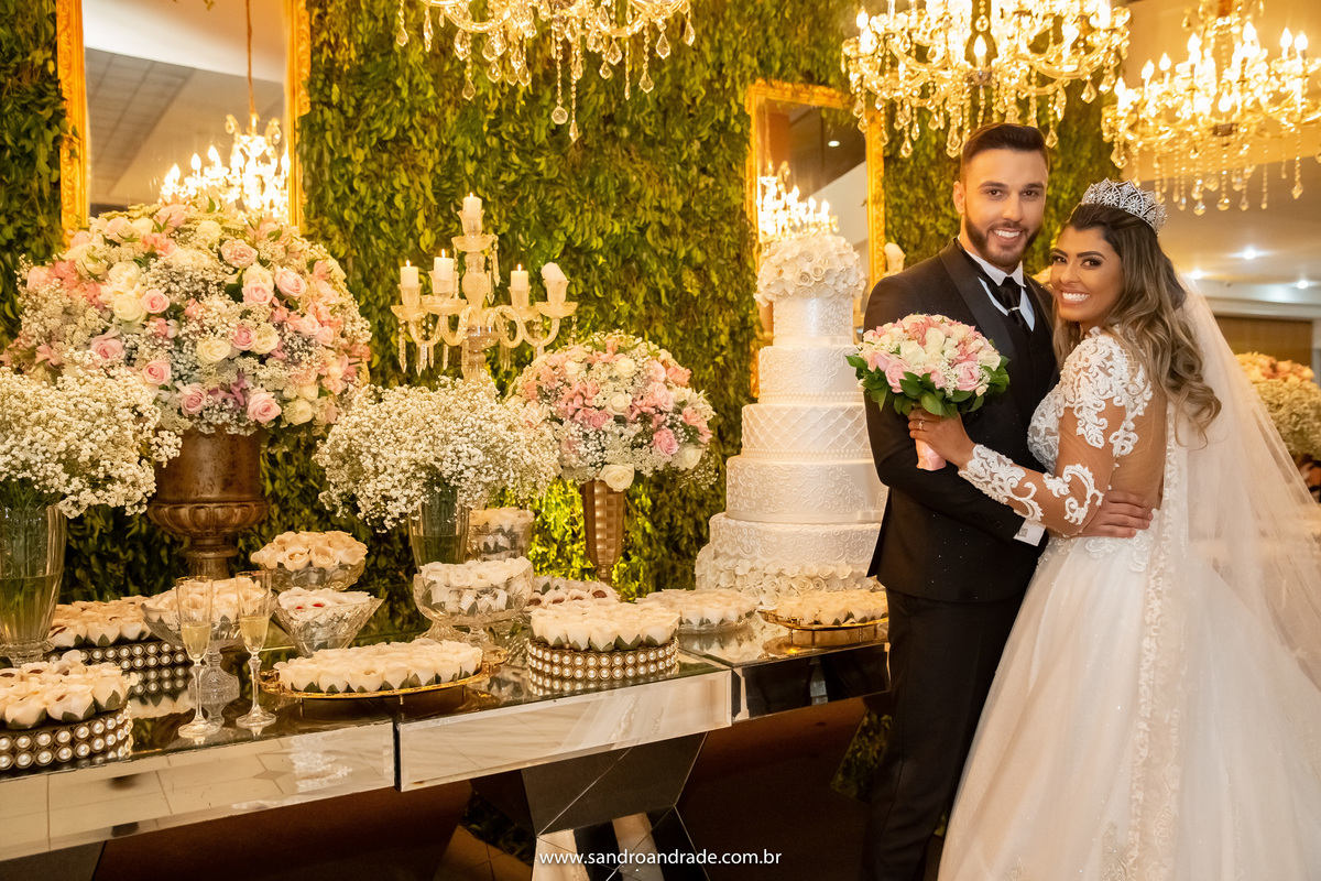 De um ângulo diferente, os noivos olham para as lentes de se Sandro seu fotógrafo e sorriem, ao fundo a mesa do bolo em detalhes.