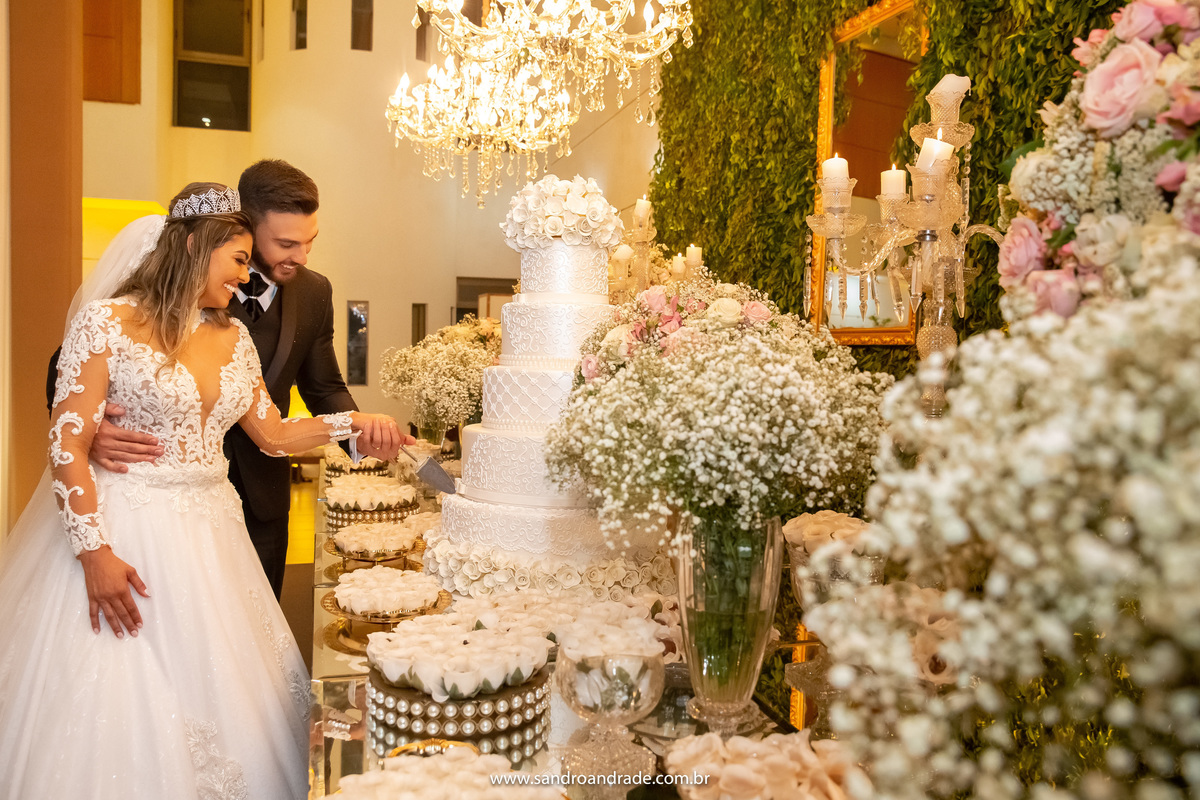 O corte do bolo, o casal está olhando para o bolo e sorrindo fazem o corte.