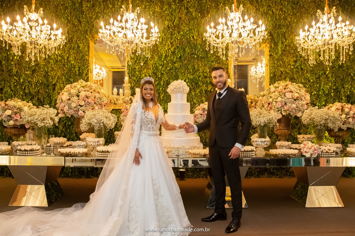 Uma foto clássica com o casal de mãos dadas na frente da mesa do bolo e doces, com o bolo ao meio.