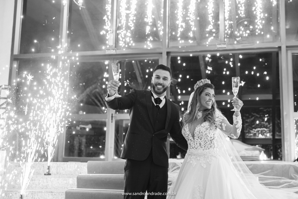 Em preto e branco, retrato do casal com o brinde durante a queima de fogos.