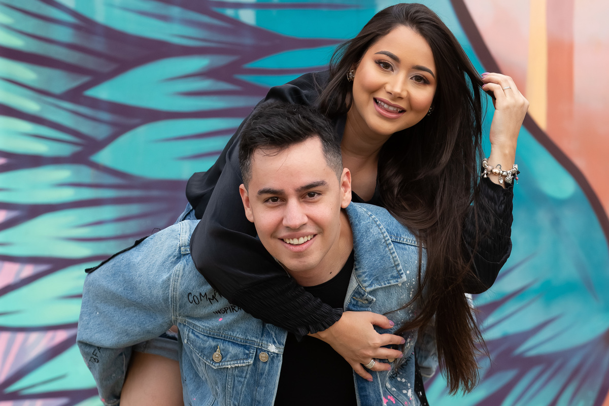 A noiva está na cacunda de seu noivo, ambos felizes e sorrindo, em uma linda imagem colorida.