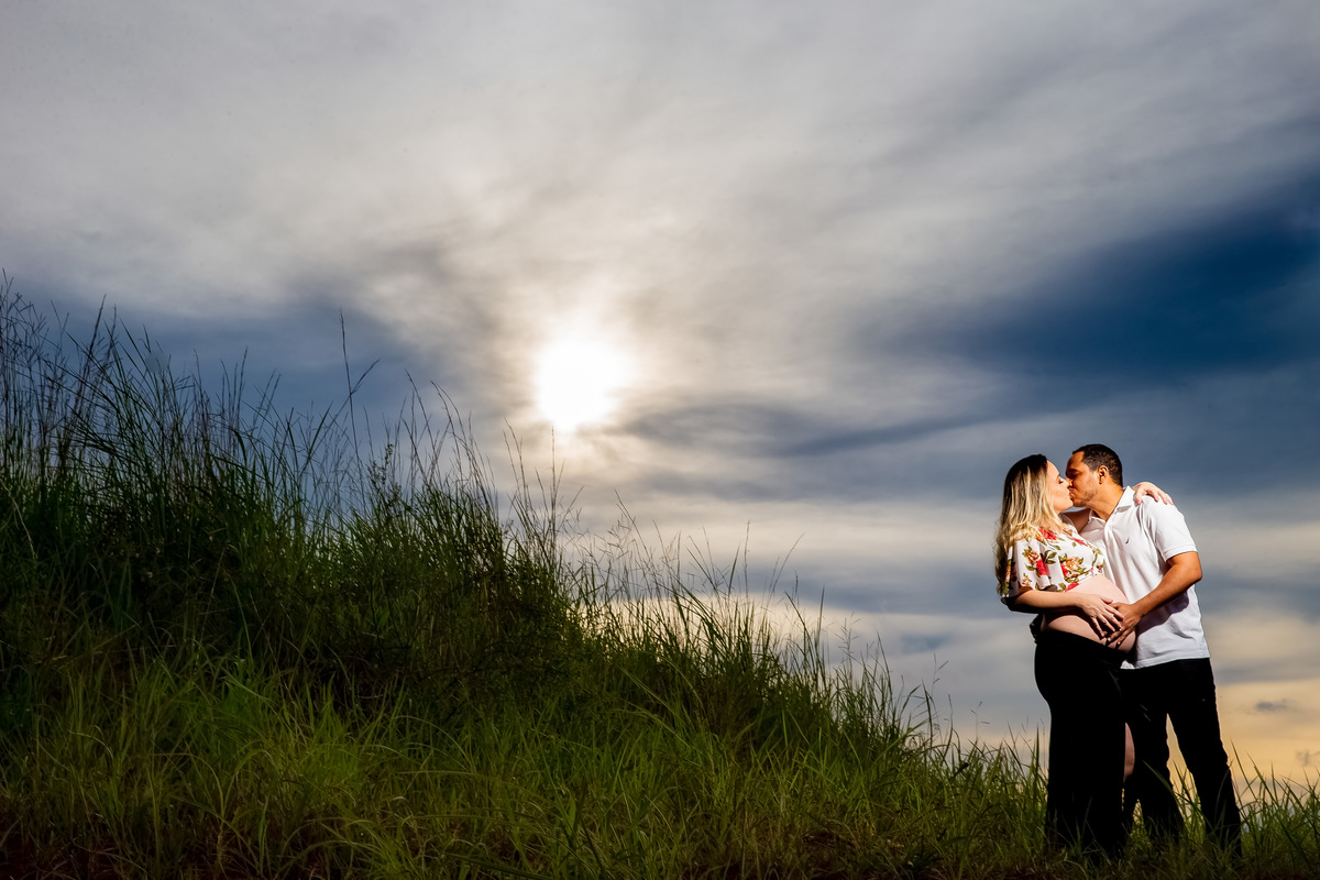 Um beijo do casal no matagal e com o céu azul cheio de nuvens, o sol escondido e muito amor envolvido na gestação.
