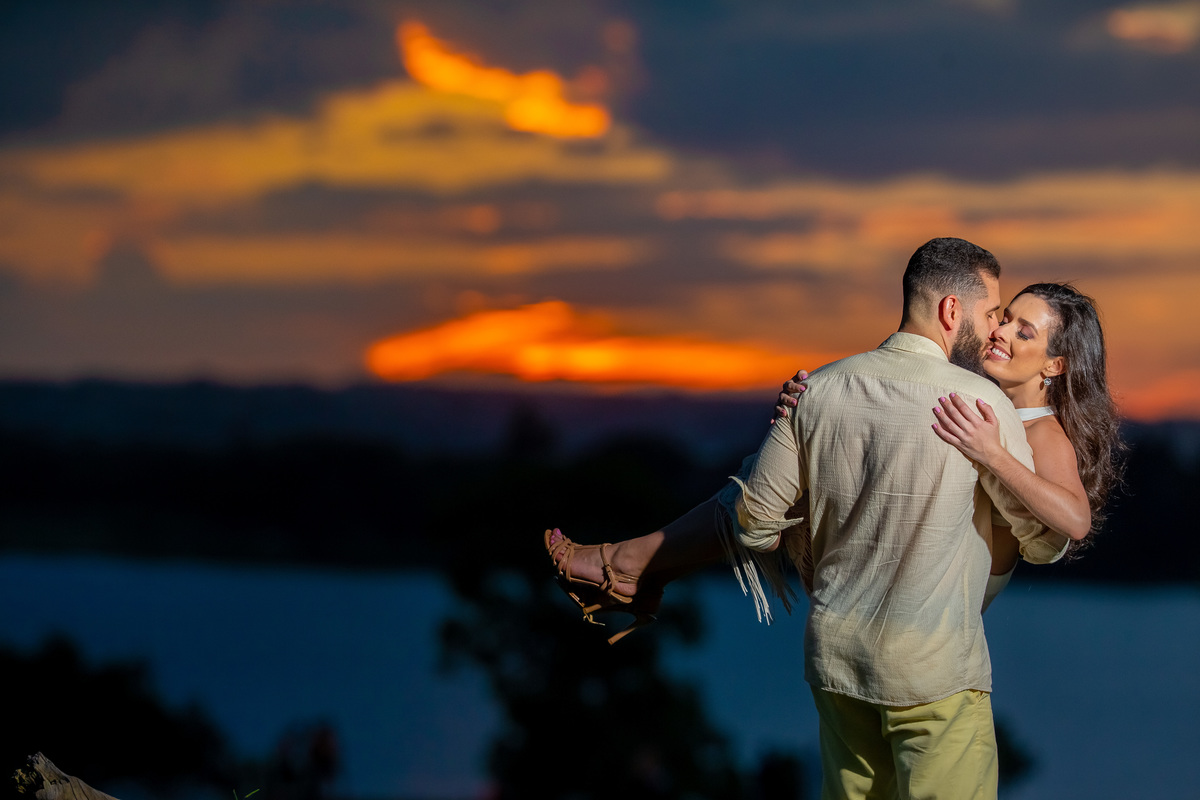 Um dos melhores fotógrafos de casamento de BSB, Sandro Andrade fez Rodrigo pegar sua noiva no colo e pediu um beijinho no canto da boca dela, um lindo retrato do casal, felizes e apaixonados na Ermida dom Bosco em Brasília com o lago de fundo e o céu.
