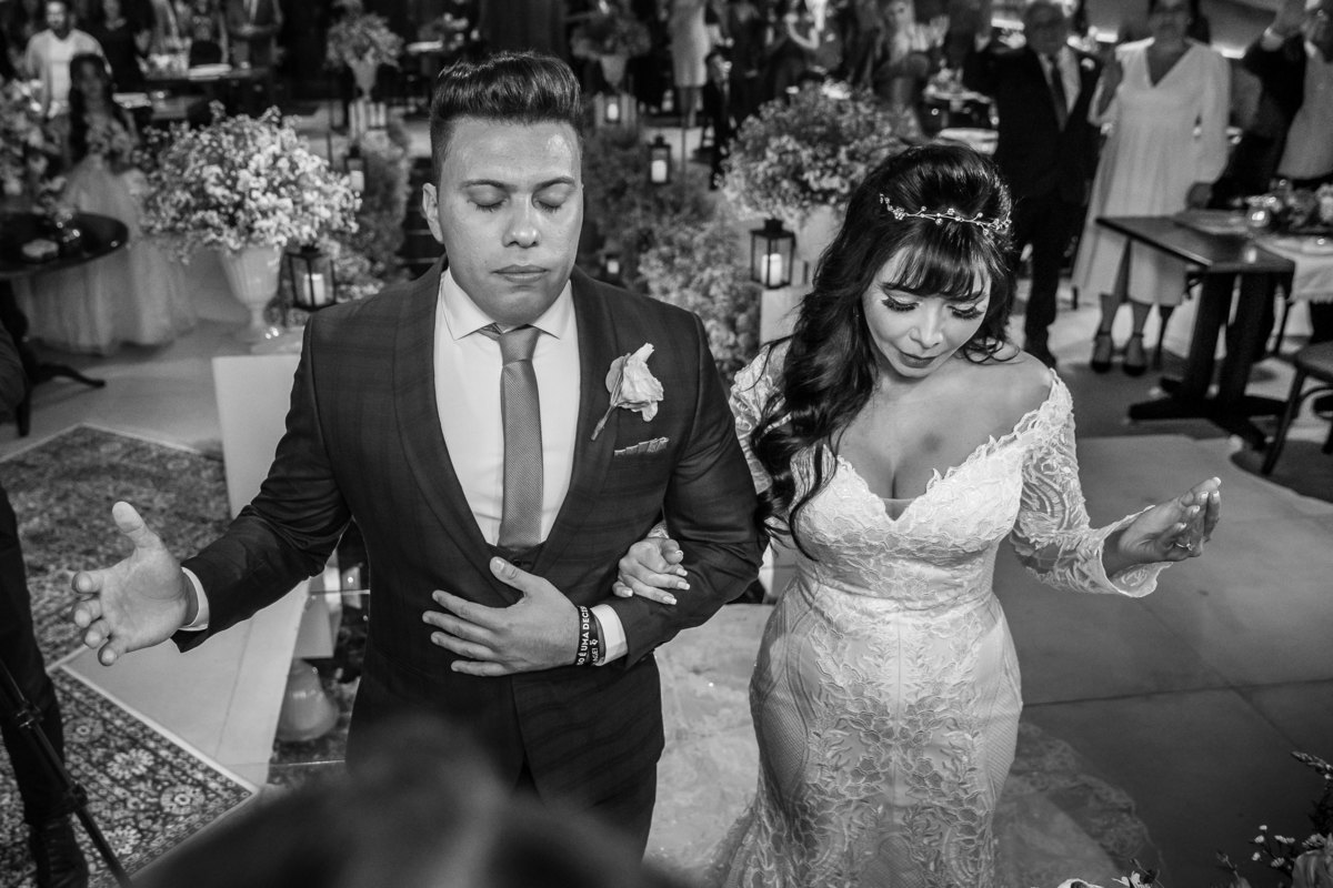 O momento da benção é sempre muito aguardado, afinal quem não deseja ter um casamento abençoado né? Cássia e Gustavo estão orando, com as mãos erguidas e de olhos fechados nesta linda fotografia preto e branco. 
