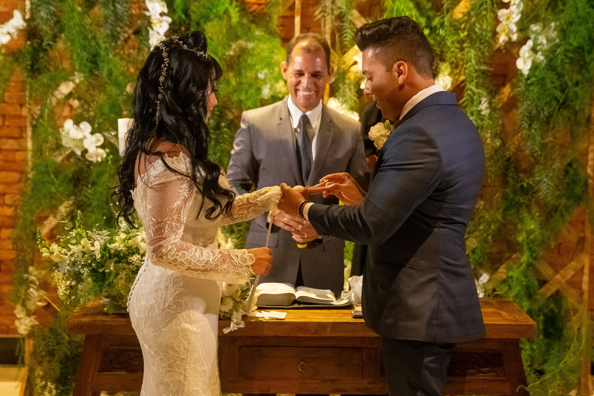 Sorrindo e tremendo de nervoso, Gustavo coloca a aliança em Cássia, uma fotografia colorida e o pastor ao fundo sorri.