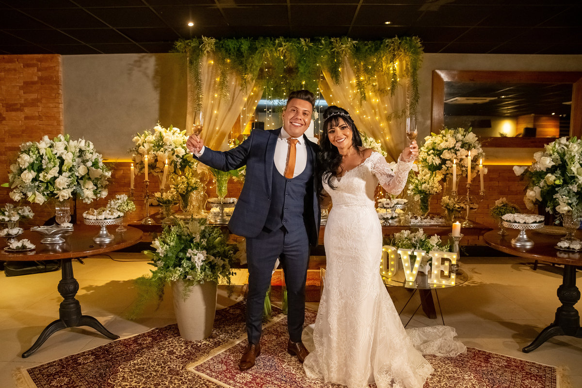 O brinde dos noivos! O casal de corpo inteiro, ergue as mãos com o brinde juntamente com seus convidados, eles estão a frente da mesa do bolo.