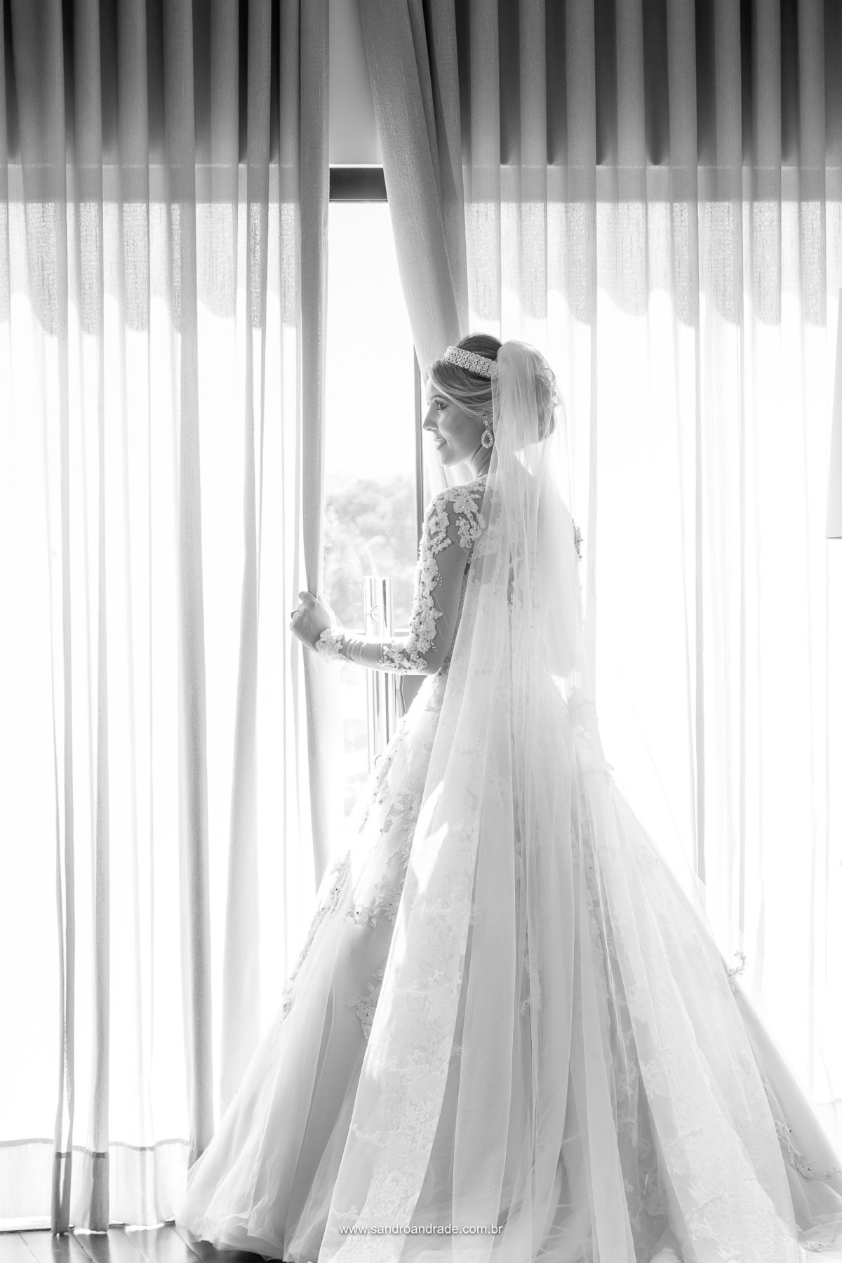 Um retrato de corpo inteiro da noiva, ela está de costas, abrindo a cortina com uma das mãos e olhando lateral sorri, feita pelo fotografo de casamento df Sandro Andrade, uma bela imagem preto e branco.