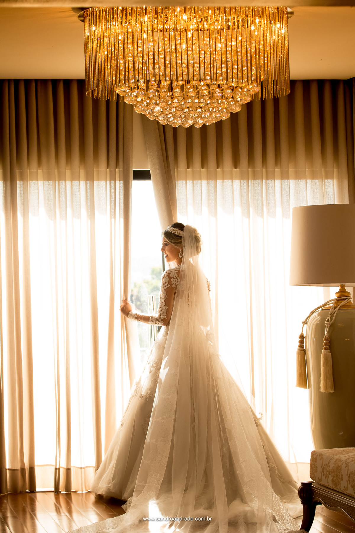 A noiva está de costas na porta do atelier Fernando Peixando, em tom amarelado, esta fotografia tem um lindo lustre no teto em composição e os móveis na lateral direita com um belo abajur.