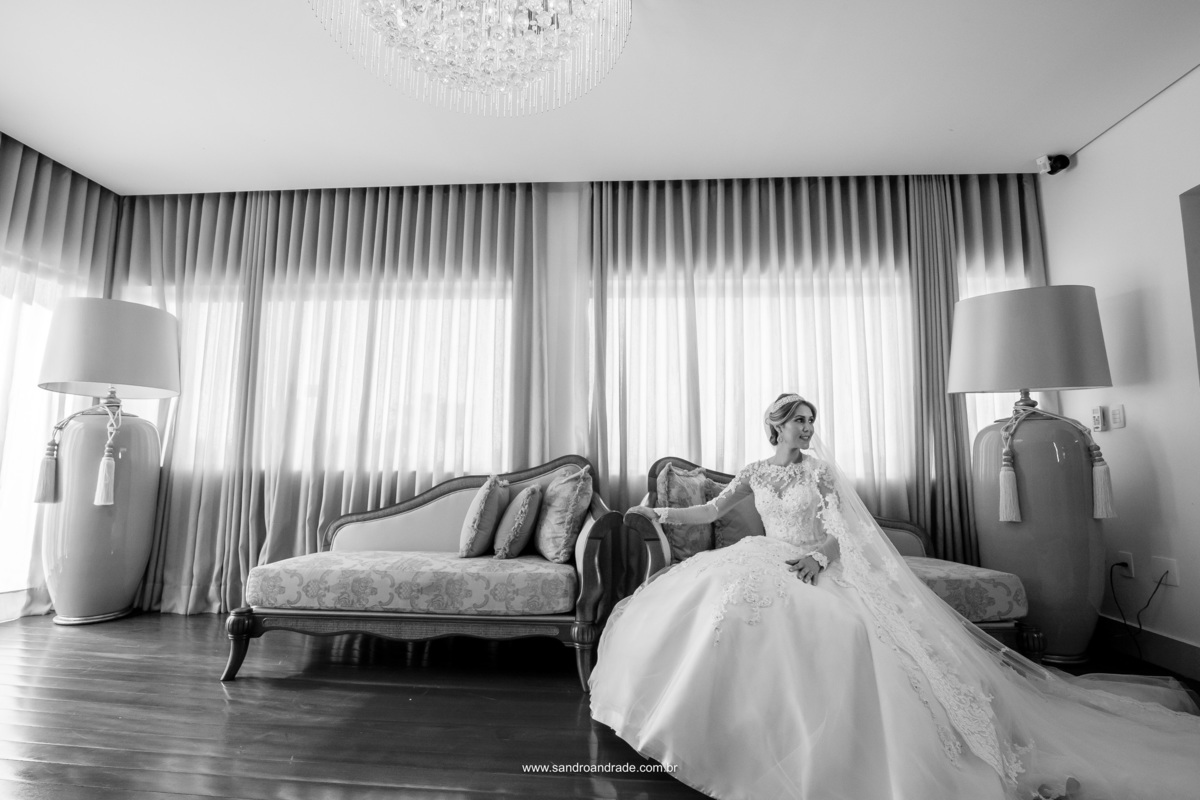 Em preto e branco, sentada no sofá estilo Luiz XV, a noiva olha para o espelho que está na lateral direita e dá um leve sorriso, com toda a sua timidez.