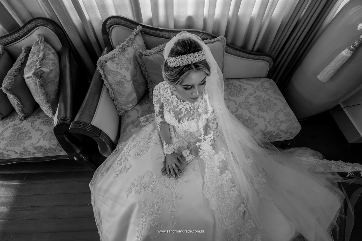 Ainda sentada no sofá, olhando lateral, Sandro Andrade faz uma foto da noiva Daiane de cima para baixo, pegando os detalhes da renda do vestido, sua tiara e maquiagem.