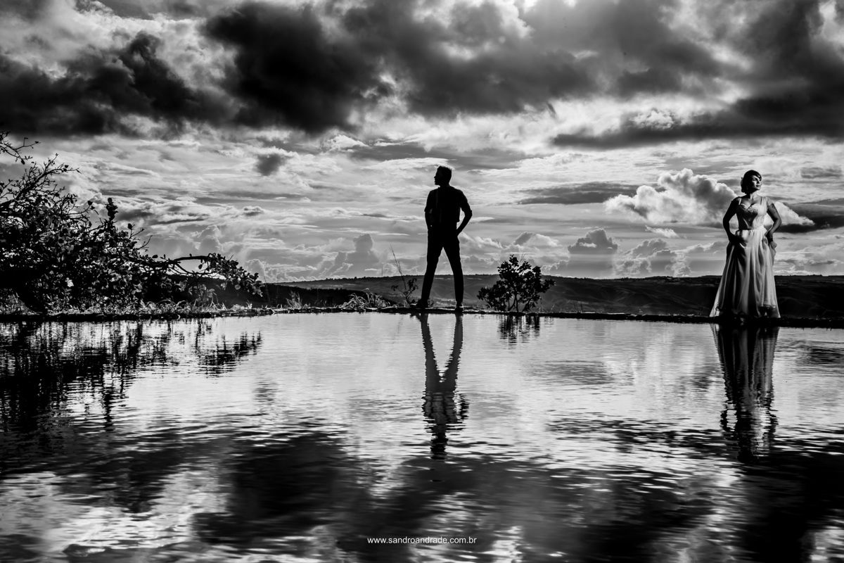 Uau...pura arte nesta linda fotografia pb. O noivo está no no centro em silhueta, segundo plano e a noiva na lateral direita iluminada com luz artificial, na esquerda uma árvore em silhueta, nuvens no céu, tudo no reflexo do lago uma belíssima composição.
