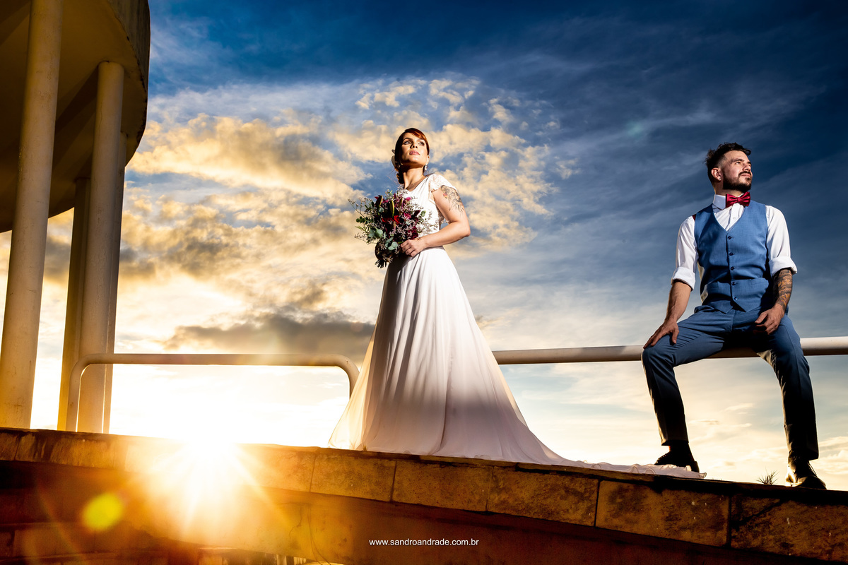 está iluminado com luz artificial, ele sentado no corrimão e ela de pé segurando seu buquê, ambos lindos e iluminados, o céu maravilindo e raios de sol que formam flairs que erradia até a parte de baixo.