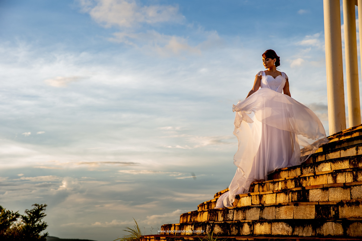No topo da escada, a noiva segura seu vestido e o vento movimenta a saia, séria, olhando para baixo, linda e serena, parece que ela está no céu.