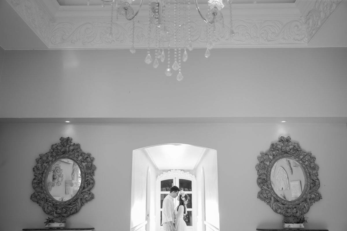 Uma linda fotografia preto e branco na entrada dos quartos dos noivos, com uma linda luz de contra, o casal no centro e em primeiro plano os espelhos e um lustre, pura arte de Sandro Andrade fotógrafo de BSB.