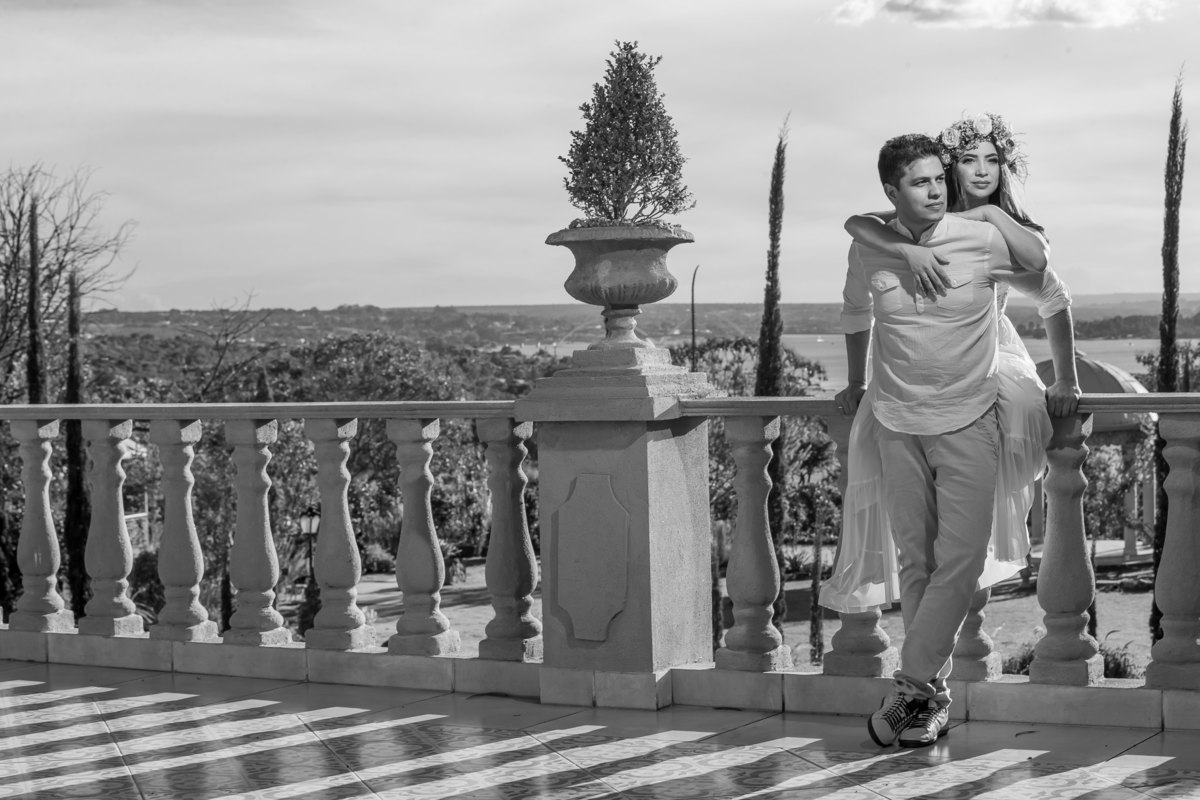 De corpo inteiro Bruce está de pé e Jessy está sentada na sacada da frente do palazzo, sérios eles olham lateral e debruçada sobre seu Bruce, ela o abraça.