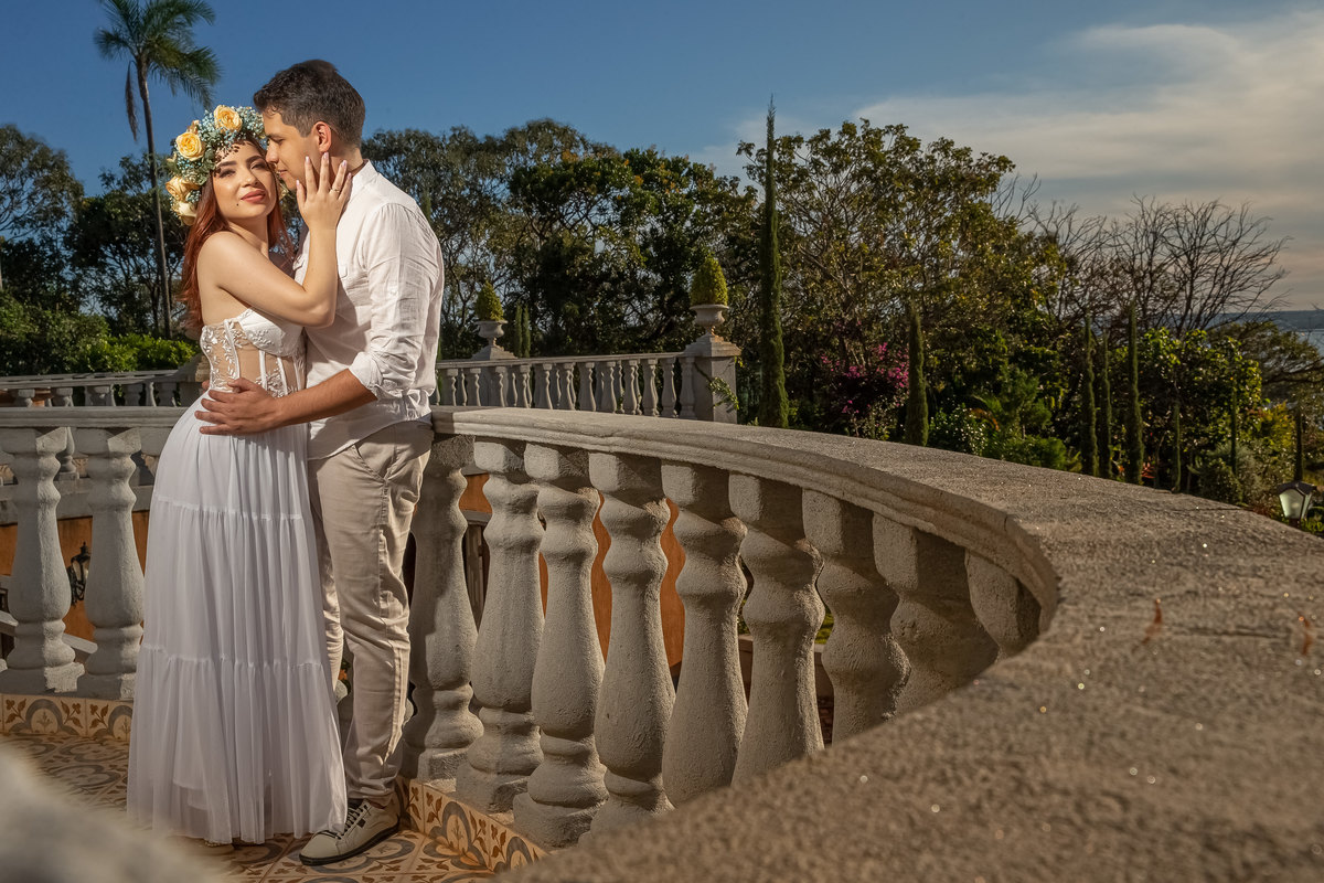 Bem juntinhos de corpo inteiro, com uma coroa de flores na cabeça, Jessy põe a mão no rosto de Bruce, em uma fotografia colorida, um lindo céu azul e toda a vegetação da Villa.