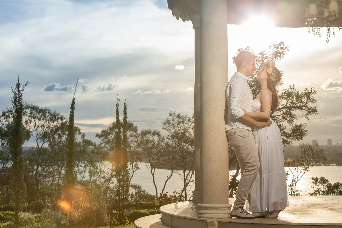 Abraçados dentro do gazebo, eles estão encostados na coluna e com um lindo pôr-do-sol ao fundo, em uma fotografia colorida com flairs que dão um toque único neste magnifico registro.
