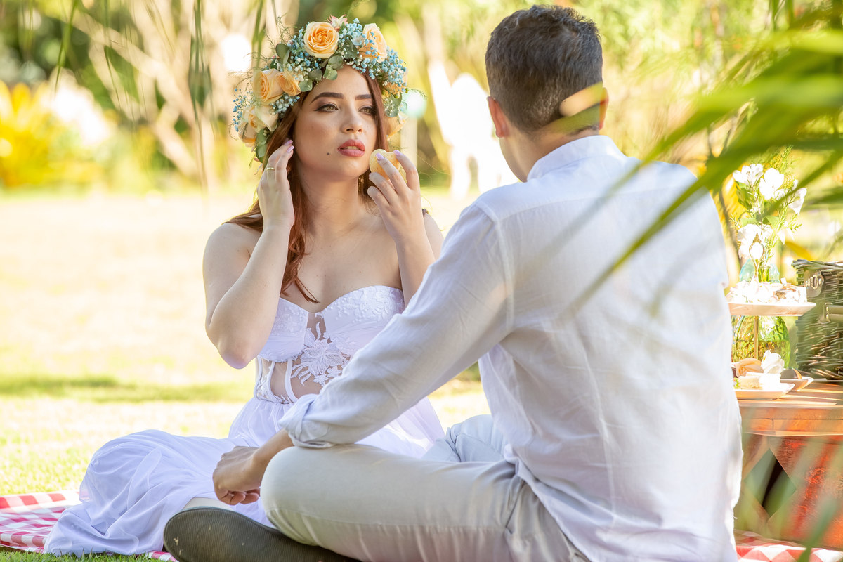 Os noivos fizeram uma pausa para um piquenique, enquanto eles aproveitam o lanchinho, uma foto do noivo de costas e a noiva de frente para ele tocando seus cabelos e com um pão de queijo em forma de coração na outra mão.