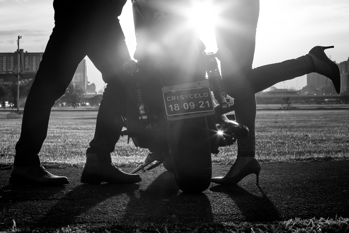 Um detalhe em silhueta, o pôr do sol, as pernas do noivo, da noiva que está com o pezinho no ar, flair e a parte da moto com a placa do casal.