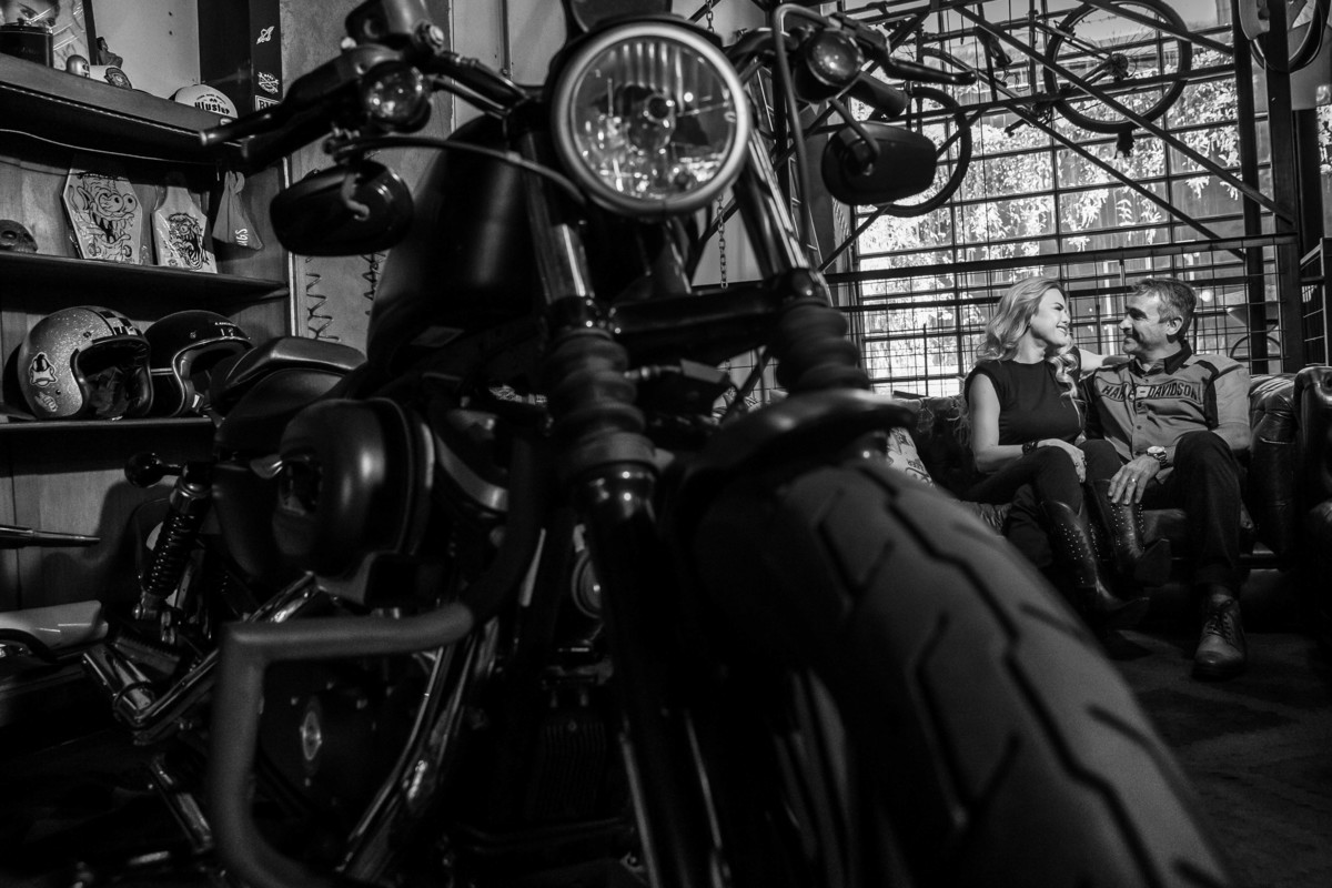 Uma linda pintura em preto e branco, a moto em primeiro plano e no canto direito da fotografia, o casal está sentado no sofá da Kluster Moto batendo papo e dando altas risadas.