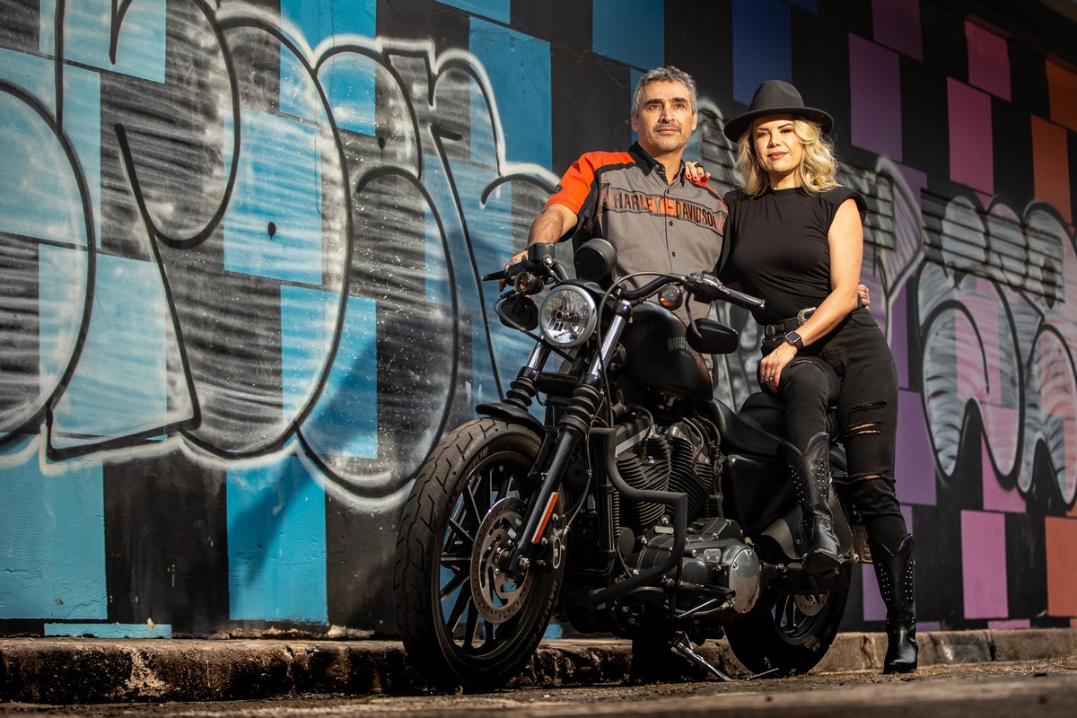 Minha nossa, cada hora que passa fica ainda melhor...com grafite na parede de fundo, o casal está encostado na moto, ele com a camisa da Harley davidson e ela toda de preto com um chapéu preto.