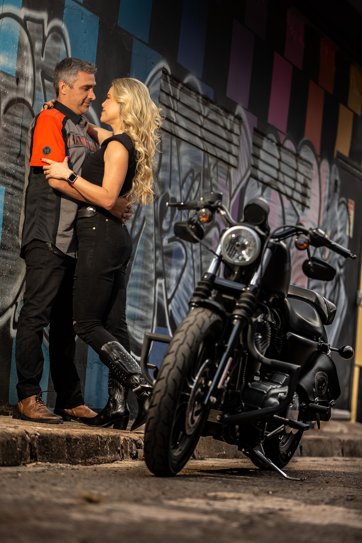 Mais uma na vertical, nesta o detalhe das botas deles e o casal se olhando apaixonados e a moto ao lado.