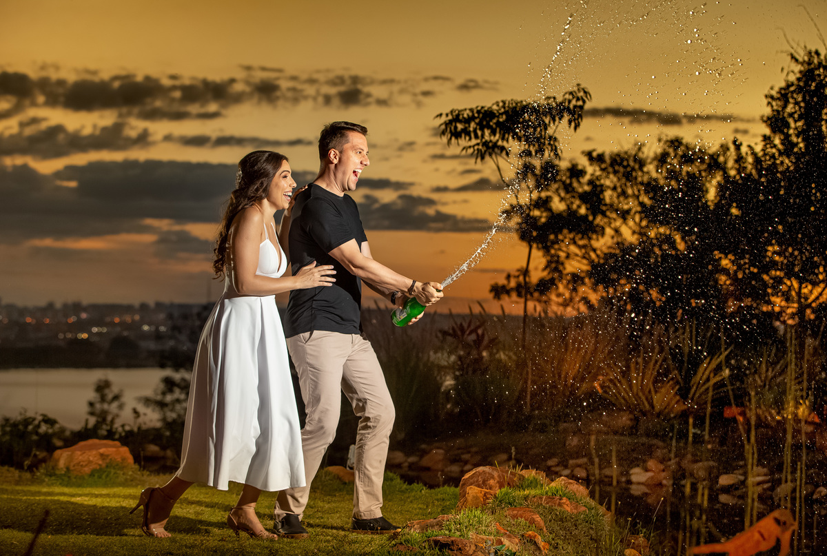Uma fotografia espontânea, o noivo está estourando um champagne, sorrindo demais e Milena está abraçando por trás dele, ambos sorriem nesta linda foto colorida.