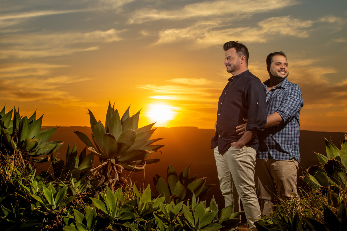 Com um lindo pôr do sol ao fundo, o céu em degradê azul, amarelo e laranja, muita vegetação do serrado e o querido casal Victor e Jorge estão abraçados.