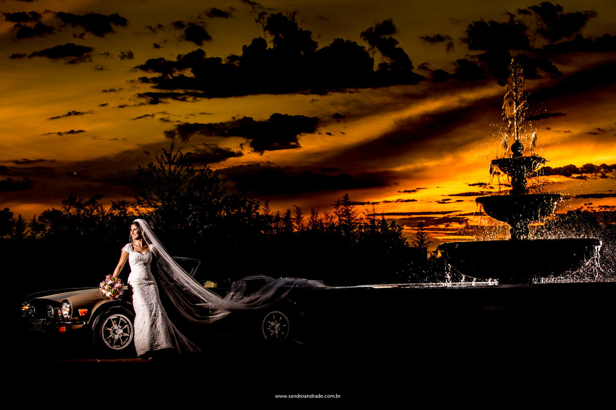 Linda fotografia feita por Sandro Andrade, num pôr-do-sol, uma linda noiva, seu carro e uma fonte contornada por luz.