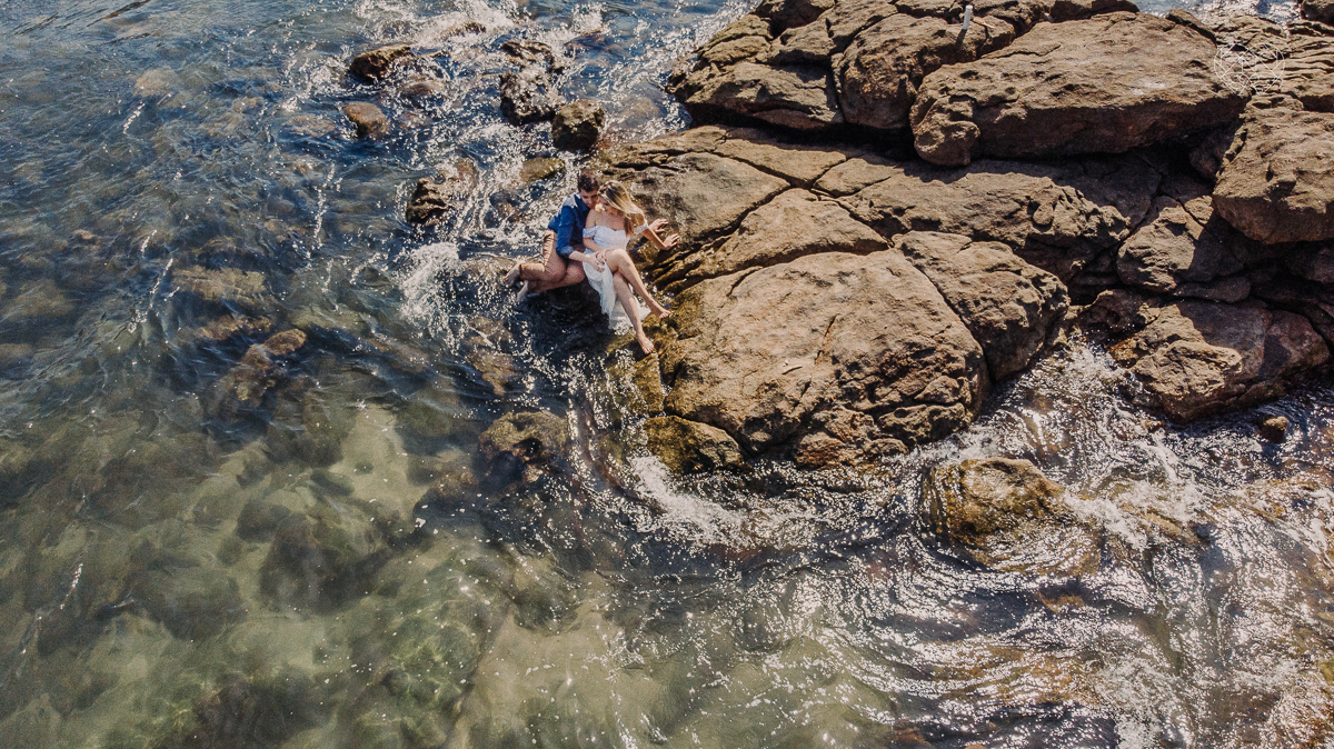 ensaio pre casamento pre wedding na praia das conchas no guaruja noiva de roupa branca e buque e noivo com roupa sociall fotos feitas na agua  na grama e correndo nas pedras ensaio feito pela fotografa premiada nayara andrade de santos