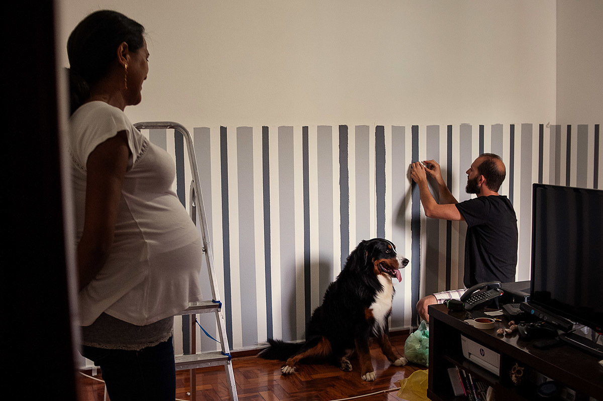 Pai arrumando o quarto do bebê, e a mãe só observa.
Cachorro olha atentamente o que seu dono está fazendo
