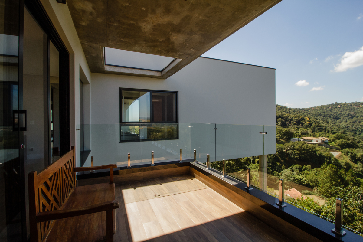 #ParaCegoVer: Sacada com piso de madeira, banco de madeira, teto de concreto com clara boia e guarda corpo de vidro.
Ao fundo temos uma parede de cor branca em balanço com uma janela sob pilotis.