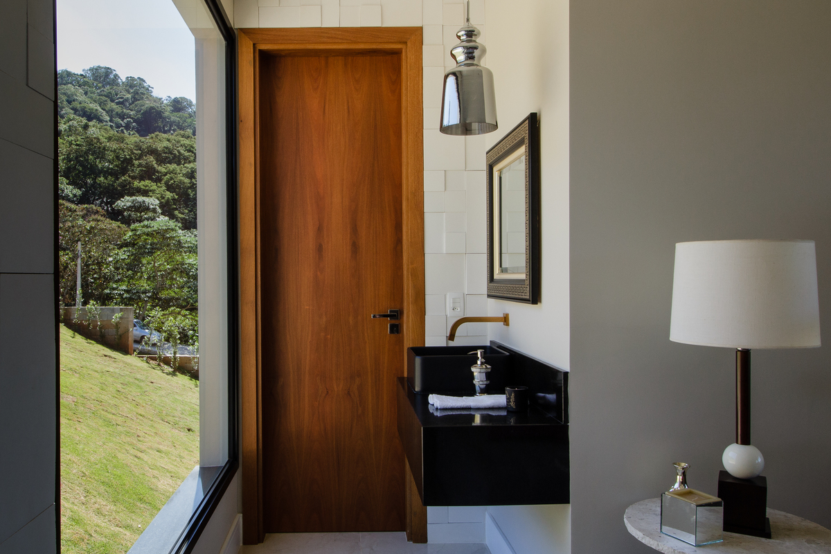 #ParaCegoVer: Fotografia do
 lavabo residência CT Quebec.
 Janela com vista para o gramado uma porta ao fundo de madeira com uma pia de pedra na cor preta, um espelho na parede e um pendente prato.
A parede possui cor bege