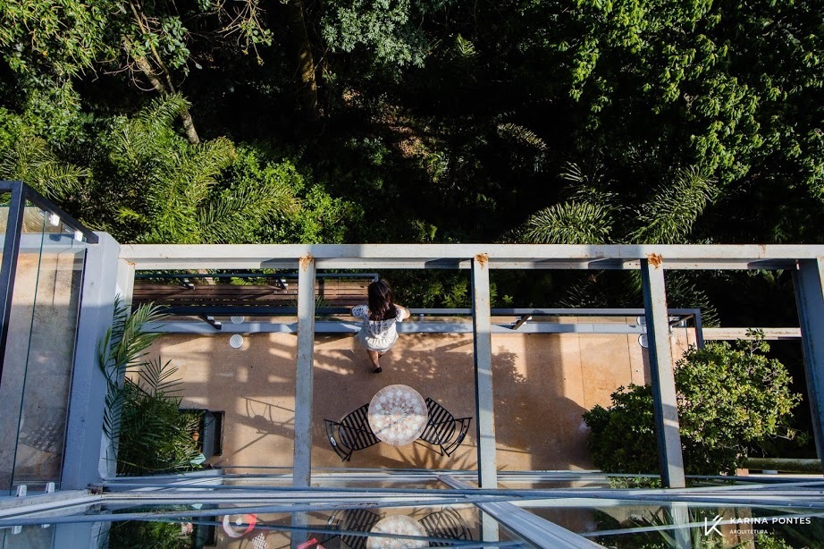 #ParaCegoVer: Fotografia da varanda vista do alto,com piso de madeira e uma mesa com duas cadeiras  e uma mulher de pé observando a mata.