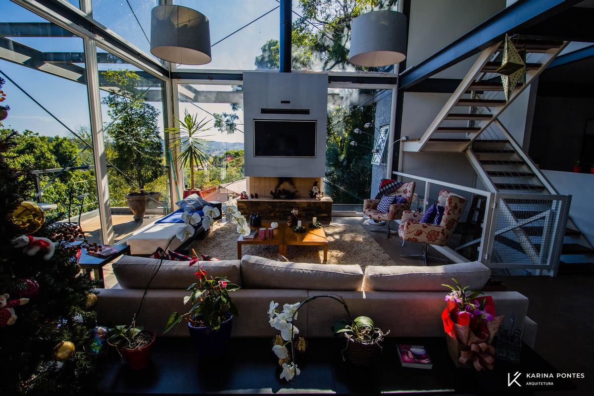 #ParaCegoVer: Fotografia colorida da sala da residência EJ Quebec com paredes de vidros com vista para mata, um aparador com objetos decorativos ao fundo um sofá branco, mesa de centro de madeira, duas poltronas colorida,painel com tv e escada branca.