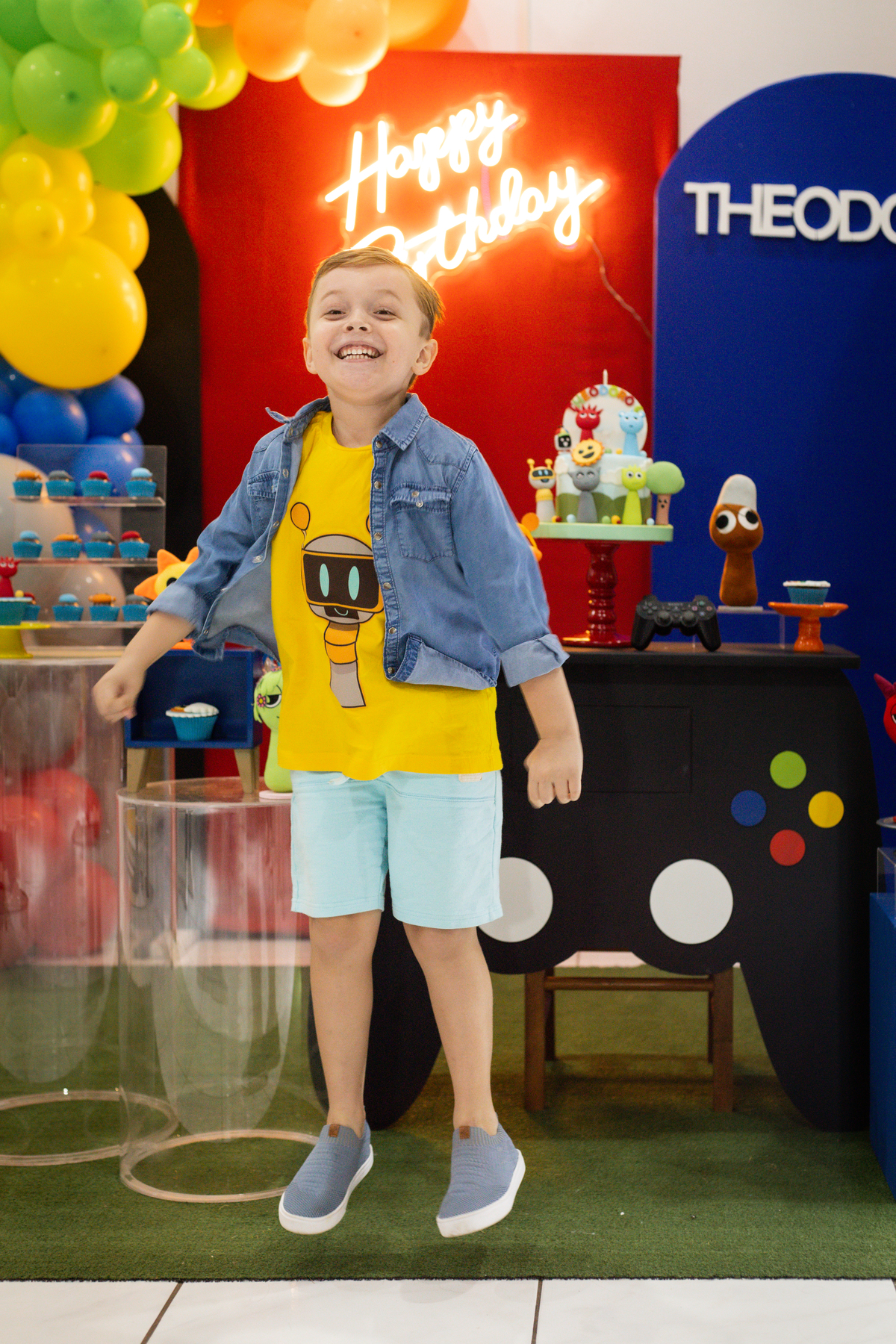 Foto do aniversariante em frente a mesa do bolo, dando um pulo no ar e sorrindo com a decoração ao fundo.