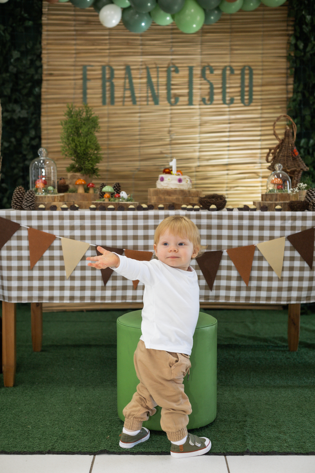 Foto do aniversariante em frente a mesa do bolo, apoiado em um puff verde virado quase de costas e jogando um beijo.