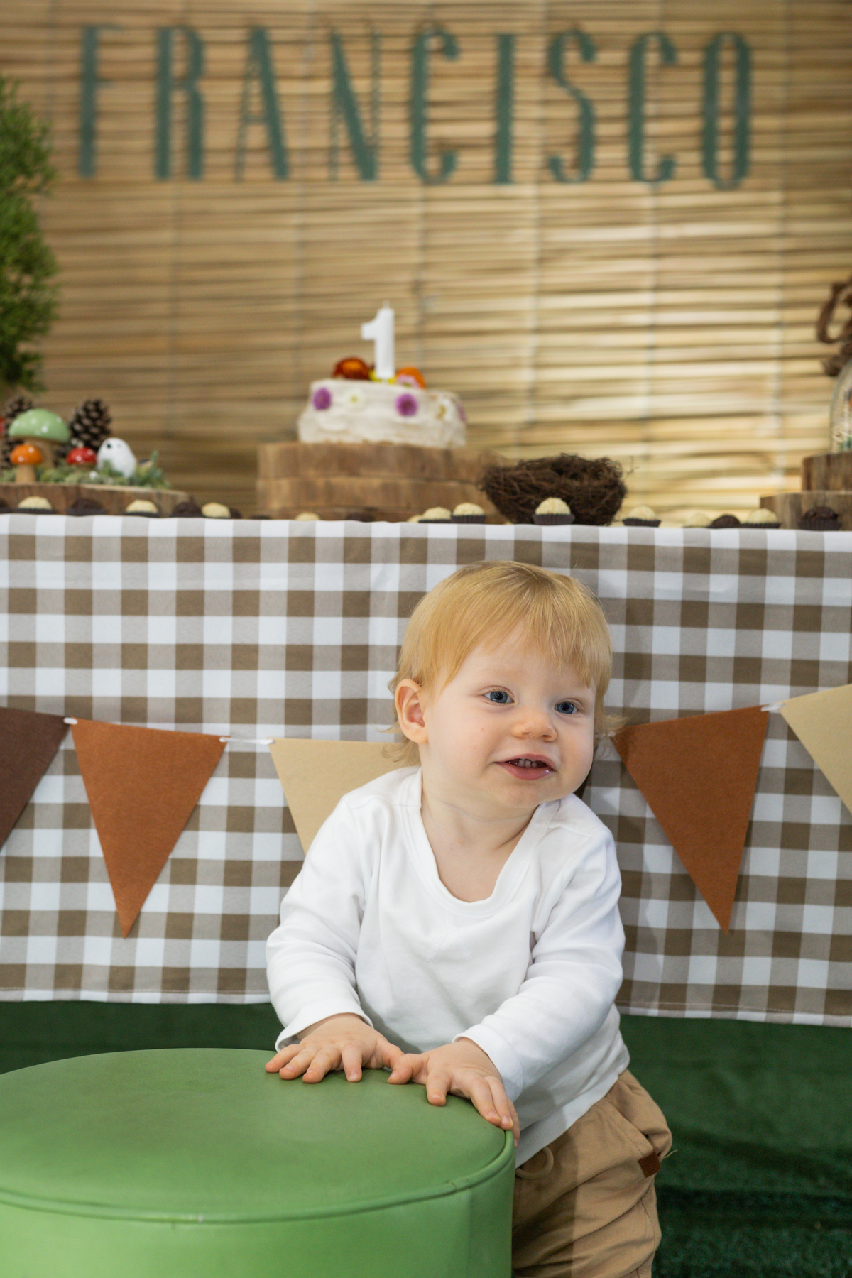 Foto do aniversariante em frente a mesa do bolo, apoiado em um puff verde, dando um sorriso.