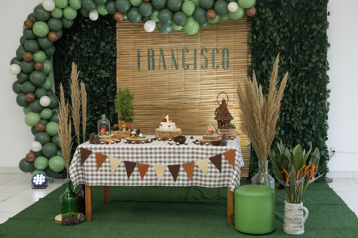Foto da decoração de festa de 1 aninho do Francisco, a imagem mostra a mesa com elementos da natureza como passarinhos, borboletas e cogumelos. Ao fundo um painel de folhas verdes nos lados com um arco de balões em tons de verde folha, no centro o nome.