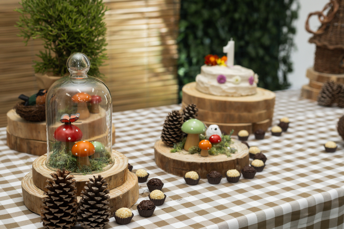 Foto de detalhe da decoração da mesa, com docinhos, pinhas cogumelos em um pequena cupula de vidro.