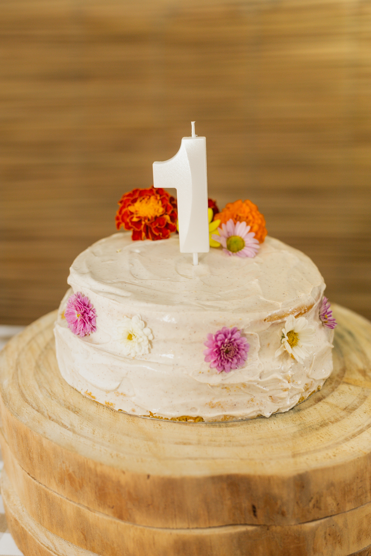 Foto do bolo de aniversário Branco e redondo, feito pela mãe do aniversáriante e decorado com flores do campo. Com uma vela de número 1 em cima.
