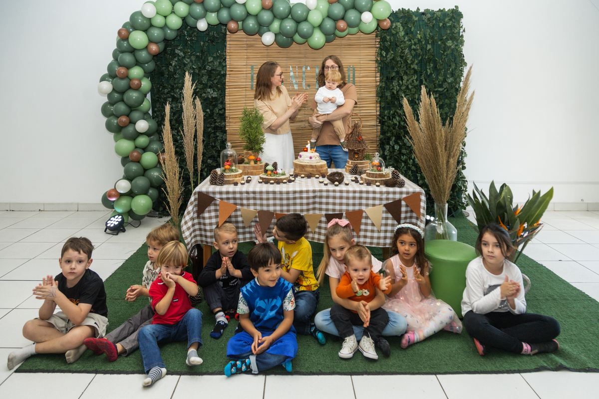 Foto do aniversariante no colo dos pais ouvindo o parabéns, sentados em frente a mesa estão as crianças presentes na festa.